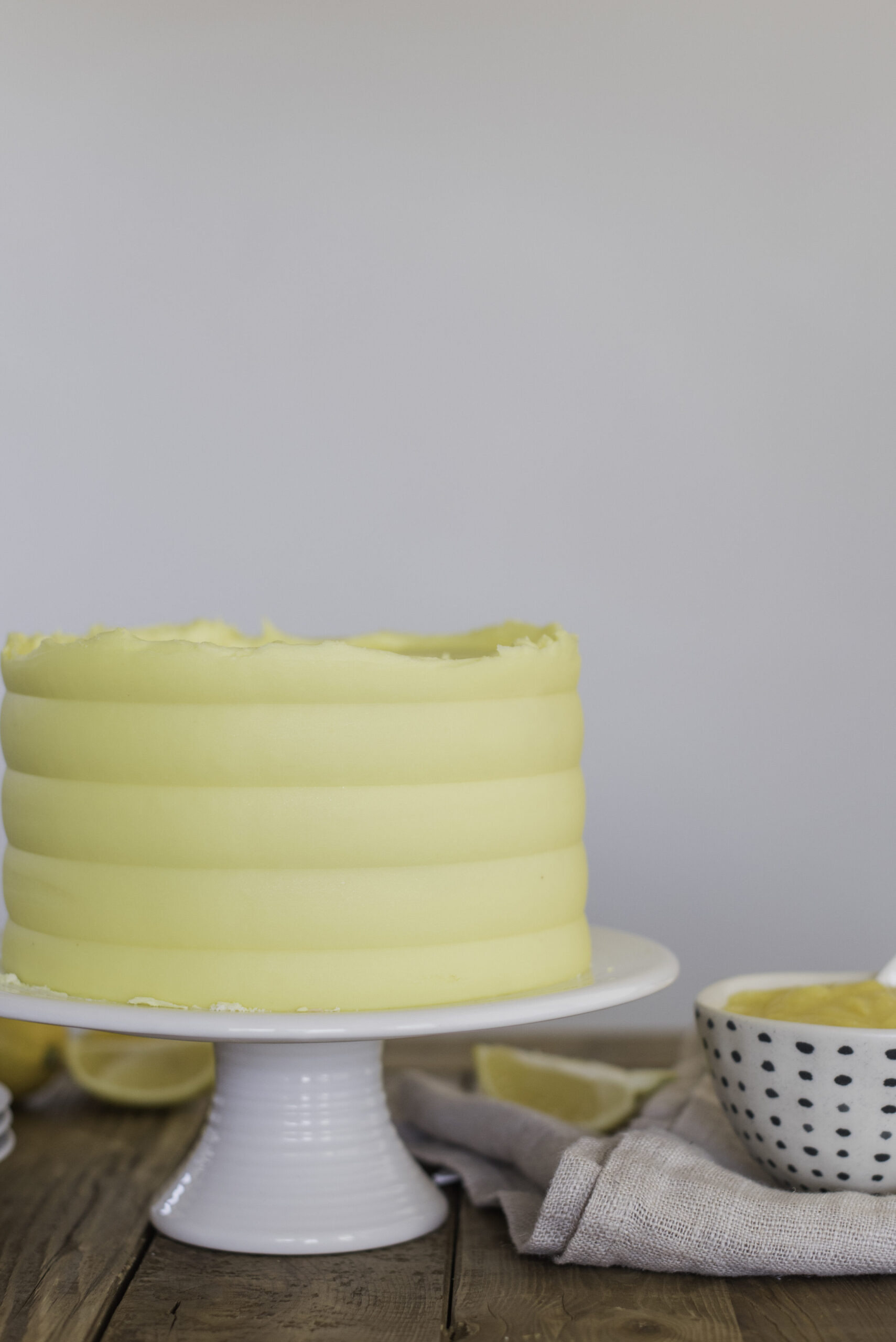 A lemon cake on a white cake stand with lemons on the table.